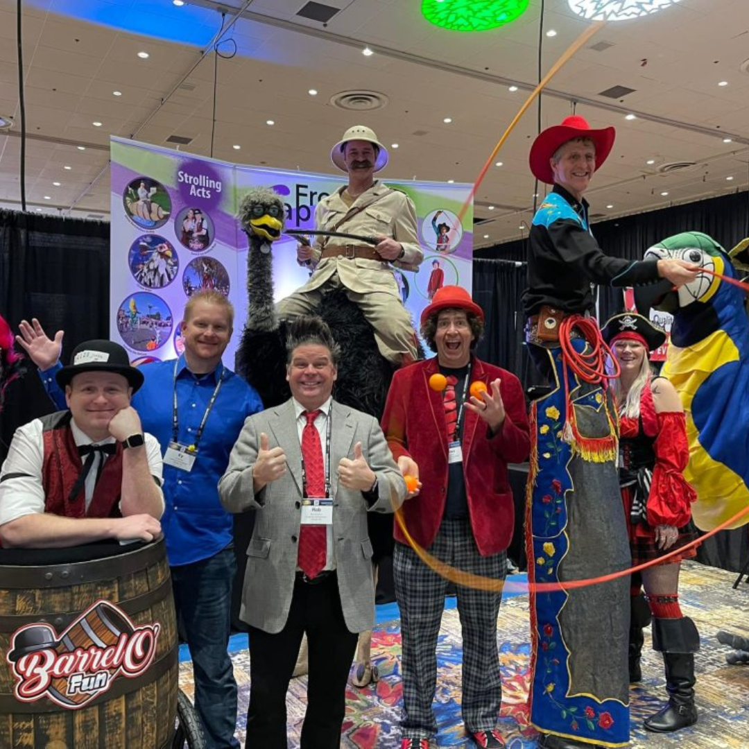 Marcus Wilson, in a red cap and jacket, juggles orange balls amid costumed entertainers, including stilt walkers, pirates, and a cowboy, at a lively event.